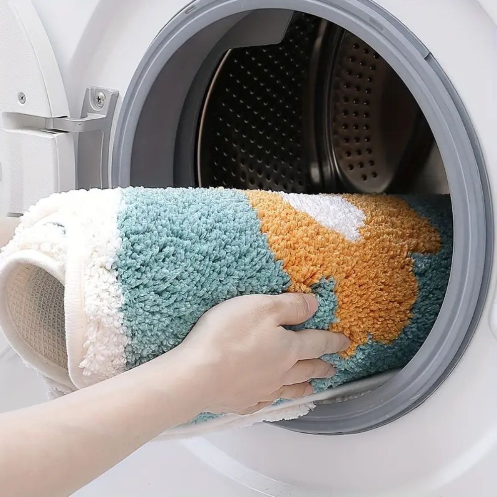 Hand placing a colorful bath mat into a washing machine.