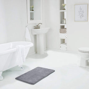 Gray bath mat on a white bathroom floor with a bathtub, sink, and toilet in the background.