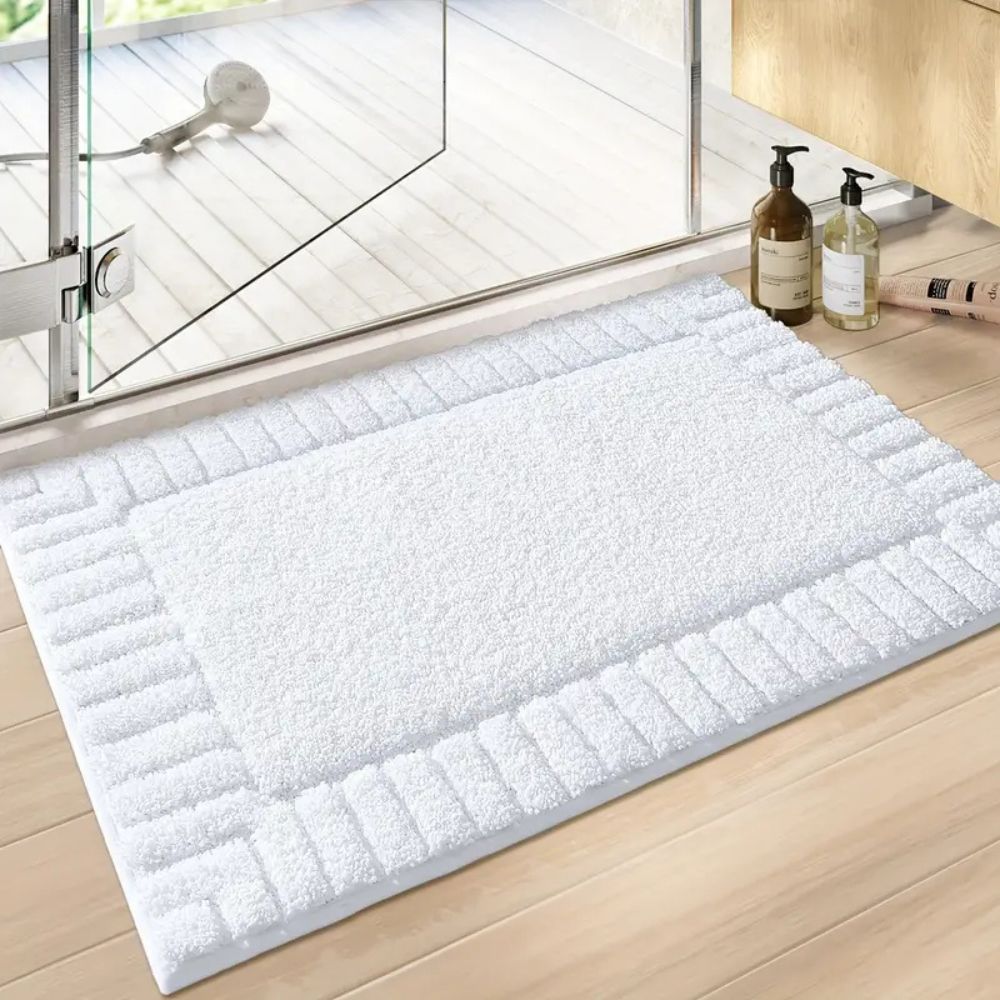 White bath mat on a wooden floor with a glass shower door in the background